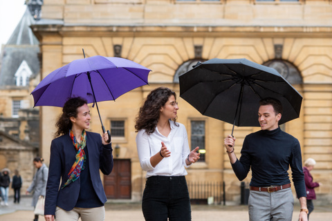 Three students walking outside with the Rhodes House building in the background. Two of them have umbrellas.