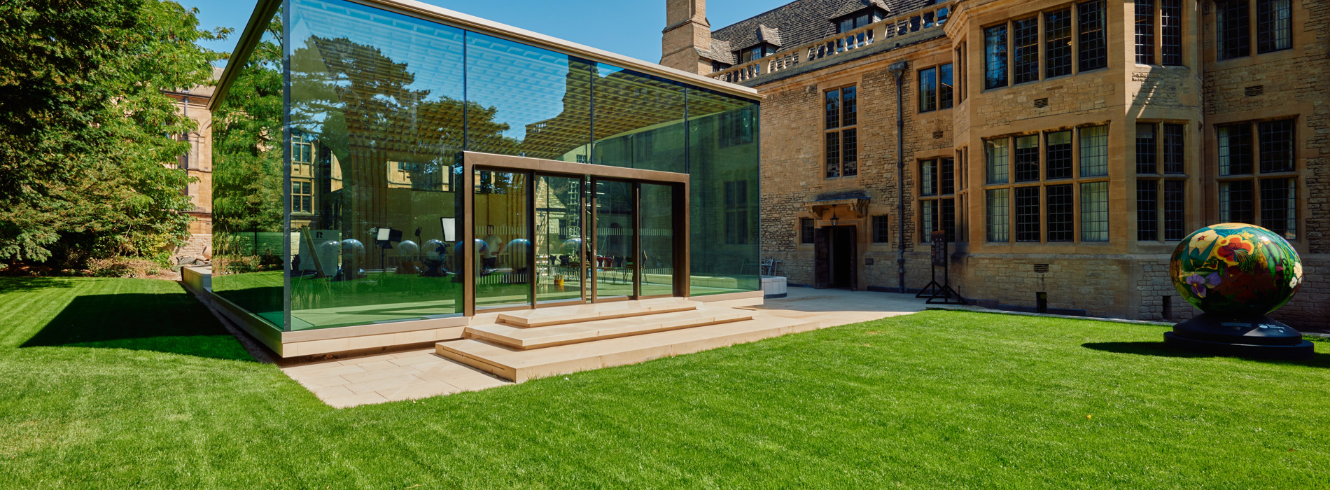 A view of the Glass Pavilion, lawn, and west wall of Rhodes House in bright sunshine.